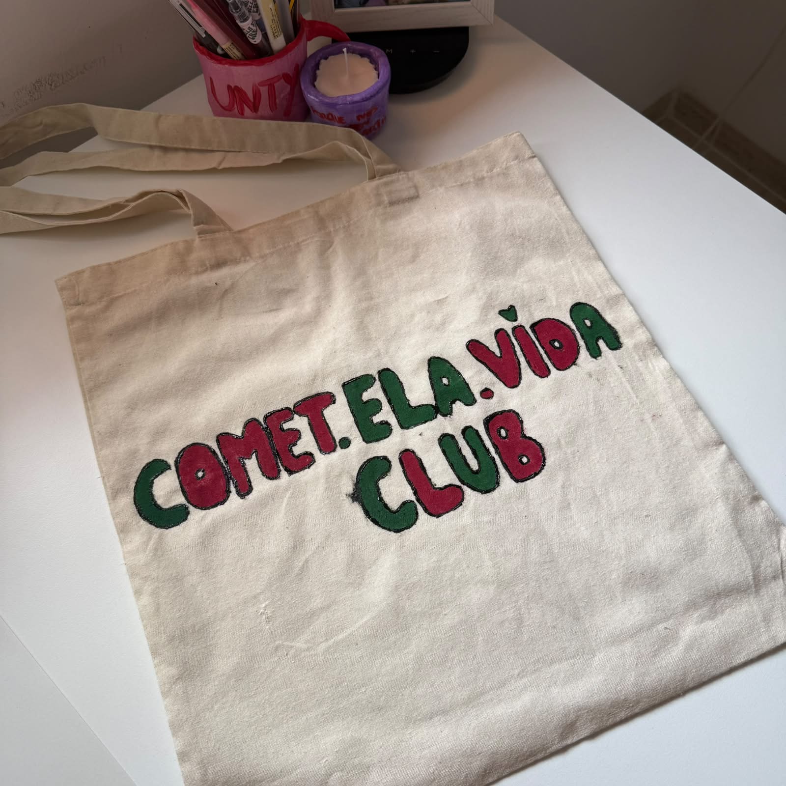 Tote bag de algodón natural con 'COMET.ELA.VIDA CLUB' pintado a mano en verde y burdeos, con un corazón sobre la i